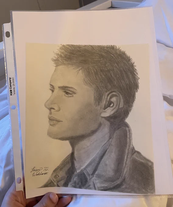 Graphite drawing of a young dean winchester's side profile. He's looking to the left of the paper and wearing a hearing aid with a leather jacket collar. Lacey's signature is in the bottom left corner.