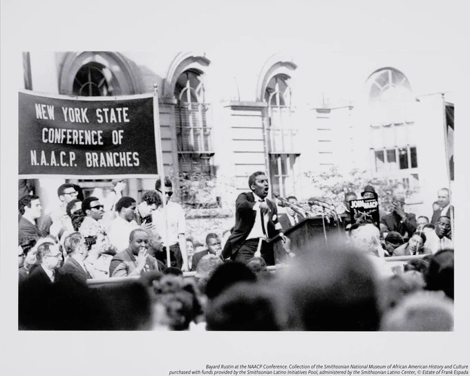 A digital print depicting a black-and-white image of Bayard Rustin standing on a stage and speaking to a crowd from a podium at the 1967 NAACP Conference in Albany, New York. Behind Rustin is a banner that reads [NEW YORK STATE / CONFERENCE OF / N.A.A.C.P. BRANCHES]. 

There are several people standing below and by the banner, on the stage and behind Rustin. To the left of Rustin is a poster held up reading [JOIN NAACP] with an image of the scales of justice. The building in the background has tall, arched windows.

Rustin is at the center of the image. He is wearing a dark suit with white shirt and dark tie. The podium is set with several microphones. His proper right hand is in motion, indicating gesturing while speaking.