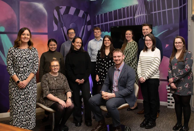Group of around 10 UNW team stood in front of purple-patterned wall smiling towards camera.