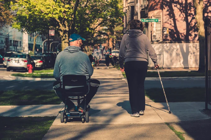 Man in blue-grey sweatshirt using power wheelchair and woman in grey hoodie using a walking stick, as they cross the road on a sunny day. Photo by @maxwbender via Unsplash.