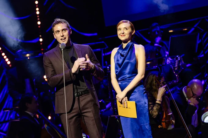 A photo showing two performers in formal clothing standing on a stage together, smiling and looking into the middle distance