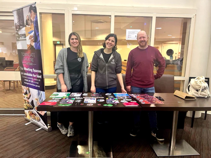 The Raspberry Pi Foundation team, Sophie, Helen, and Fergus, stood behind a table with stickers, leaflets, and posters.