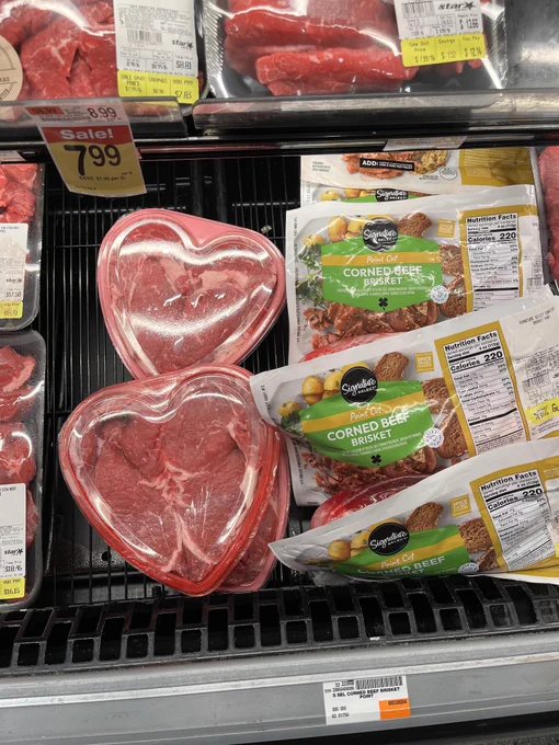 Portion of the beef section within a grocery store. Surrounded by other cuts of meat, there exists several packages of corned beef brisket, along with three red, heart-shaped plastic containers with raw beef inside.
