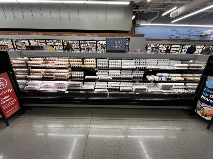 Well-stocked aisle of assorted egg cartons within a grocery store. Several people are seen shopping in the background next to other, refrigerated sections.