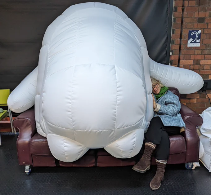 A person sitting on a sofa next to a VERY LARGE inflatable robot. The robot is white and round, it is faceless with two arms and two feet. It looks very cute and huggable.