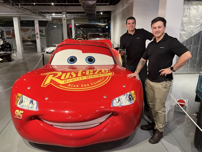 Two men standing beside a car resembling Lightning McQueen, the famous character from the movie Cars.