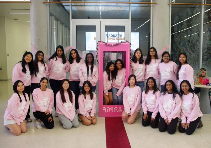 19 women wearing matching light pink sweatshirts pose for a photo inside a building. Two people in the middle are inside of a pink box that says “WCIS” at the bottom. Behind them is a room with class doors and walls, a screen can be seen inside. To the side of them, someone sits at a table on her computer with a water bottle and a can on the table.Photo courtesy of Urmi Mistry.