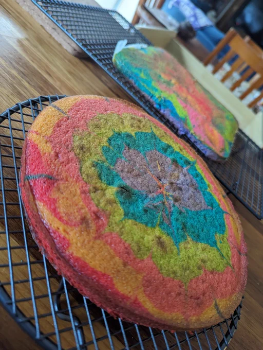 A round tie dye cake with a mixture of cascading colours, purple, blue, green, pink,red and yellow. In the background is a rectangular cake with similar colours.
