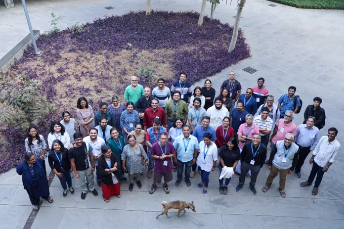 A group of educators and computing science education specialists grouped outside for a photograph taken from high up (possibly a window or roof). A dog is walking past the group as the photo is being taken.