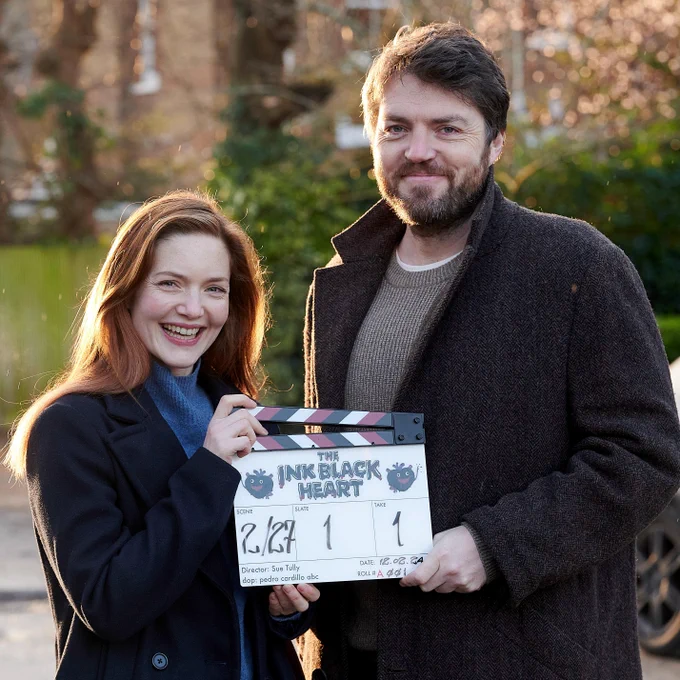 Tom Burke and Holliday Grainger outdoors behind the screens on Strike, smiling, holding a clapperboard for The Ink Black Heart