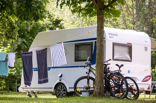 Auf dem Bild ist ein weißer Camping-Anhänger zu sehen. Davor steht ein Baum, an dem eine Wäscheleine mit Handtüchern gespannt ist. Zwei Fahrräder sind am Baum angelehnt.