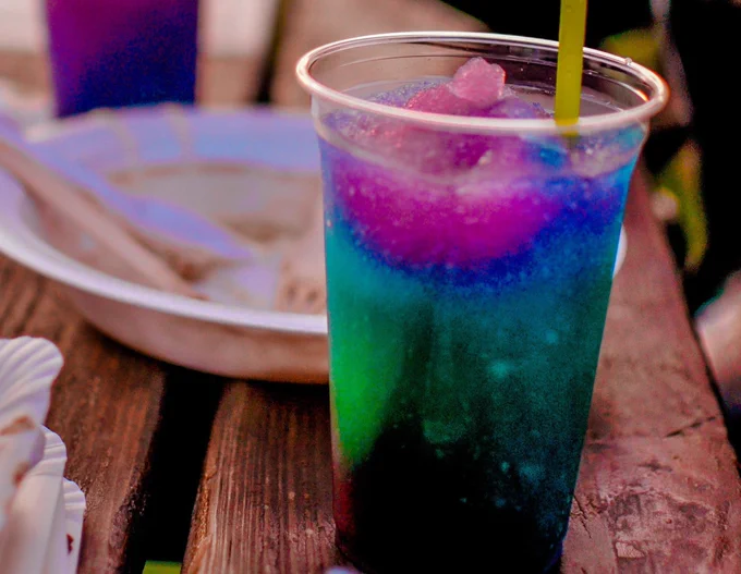 Image of brightly coloured slushy drink in plastic cup with yellow straw on wooden table