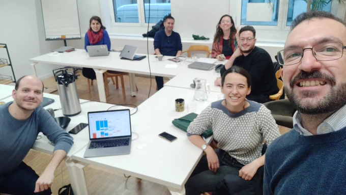 The photo shows 7 members of the IRM team dotted around a U-shaped table filled with laptops and papers. Everyone is smiling and looking at the camera.