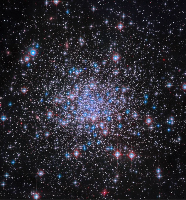 Thousands of bright stars shine against black space, more tightly condensed near the image’s center. The stars glint in shades of white, blue, and red, and diffraction spikes are visible around the foreground stars.