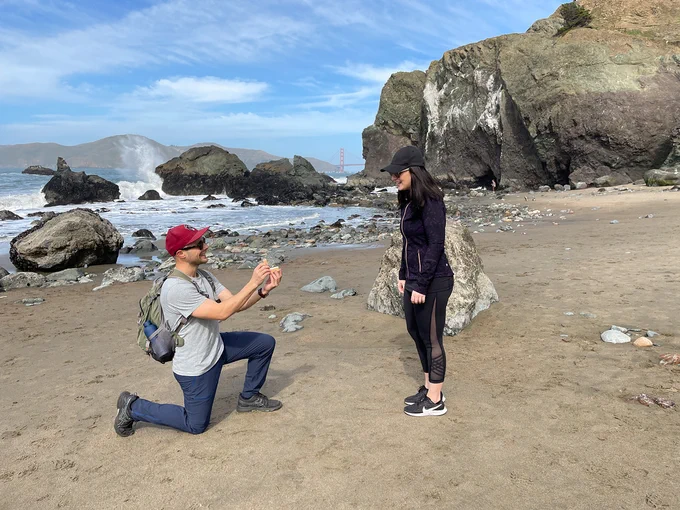 A person kneels with a ring in front of a standing person on the beach.