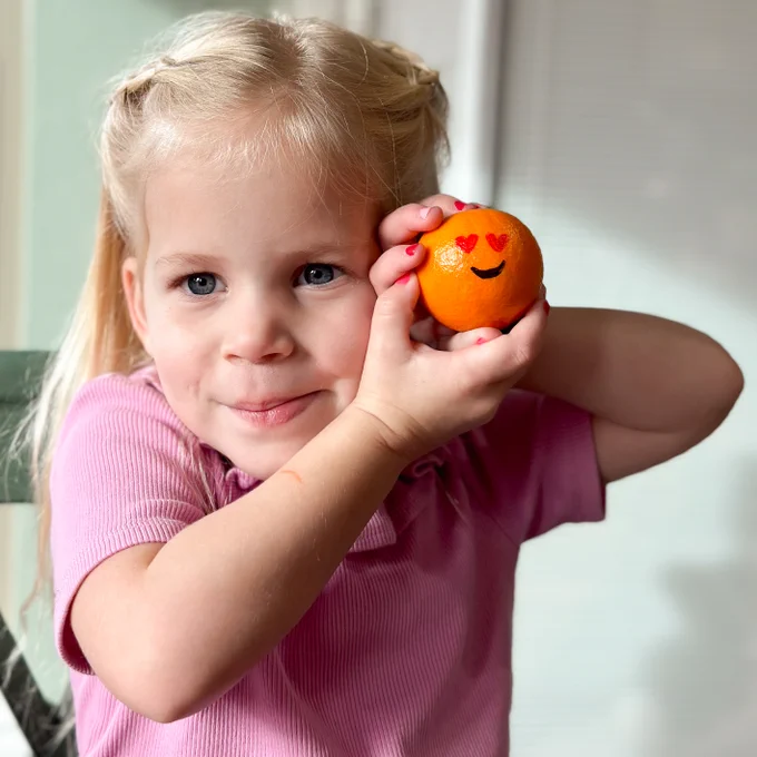 A little girl holding up a cutie with heart eyes drawn on it.