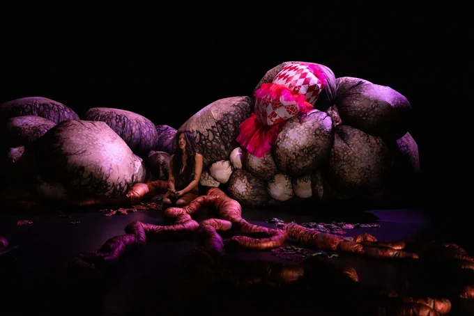 A moment from Justin Shoulder’s Anito: multicoloured fabrics, shapes and textures dwaf a person with long hair kneeling on the ground amongst them.