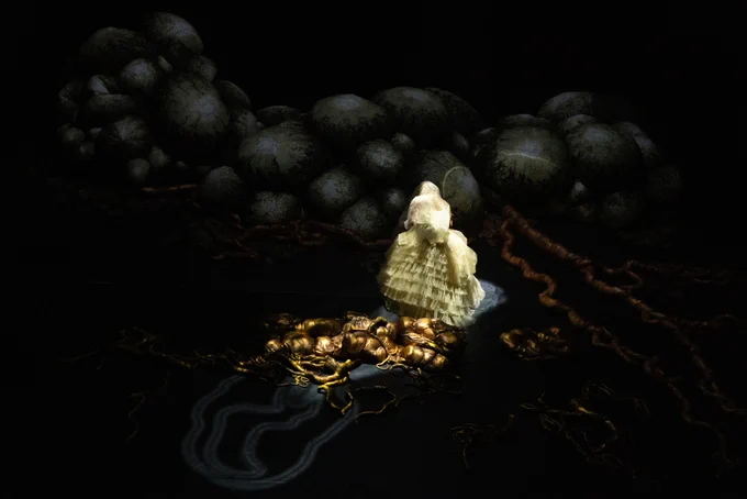 A moment from Justin Shoulder’s Anito: a figure in a white dress with a tiered full skirt faces away from the camera. Behind them lies a tangle of golden-coloured fabrics across the dark stage.
