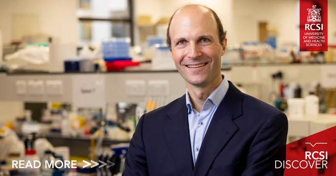 A man stands in front of a lab (the background is quite blurred). He is wearing a navy suit jacket and an open necked blue shirt. He is smiling. In the right hand upper corner the RCSI logo is highlighted in red. In the bottom right hand corner are the words 'RCSI Discover' highlighted in red also. 