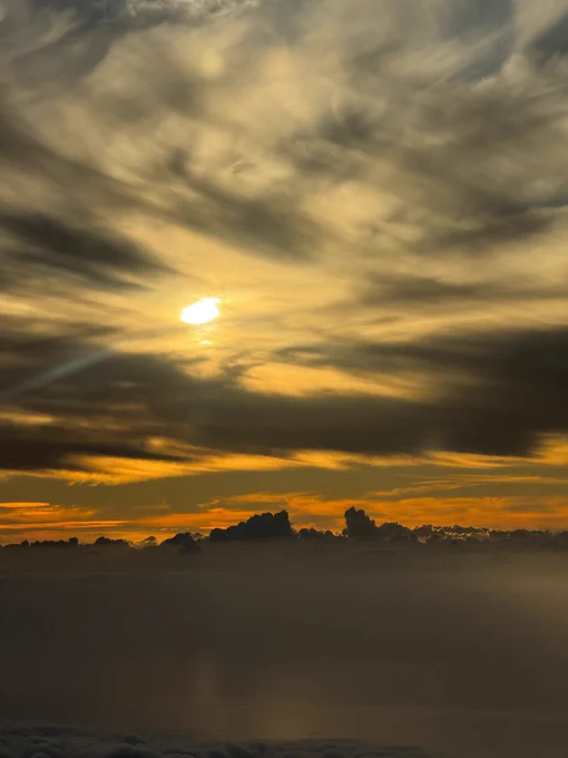 A dramatic sky with the sun partially obscured by clouds, giving off a soft, glowing light. The clouds are a mixture of dense cumulonimbus near the horizon and streaky cirrus above, all bathed in deep orange and yellow hues from the setting sun. The lower part of the image is shrouded in shadow, possibly suggesting a layer of fog or a body of water below, reflecting the sky's intense colors and contributing to the scene's moody atmosphere.