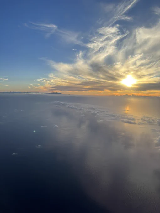 A serene view of the sun setting over a calm ocean, with the sky painted in a gradient of blues, yellows, and oranges. Wispy cirrus clouds stretch across the sky, and below, a layer of cumulus clouds hovers above the ocean, reflecting the sunset colors. The horizon is visible in the distance, and the sun's rays create a dazzling effect on the water's surface.