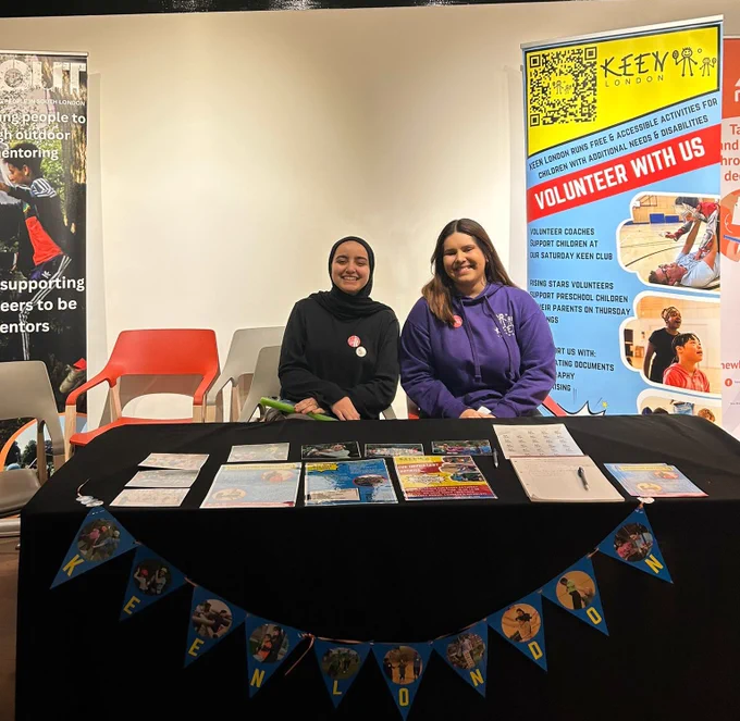 Photo of samira wearing black and Viktoria wearing a purple keen hoodie smiling next to a volunteer with us keen banner