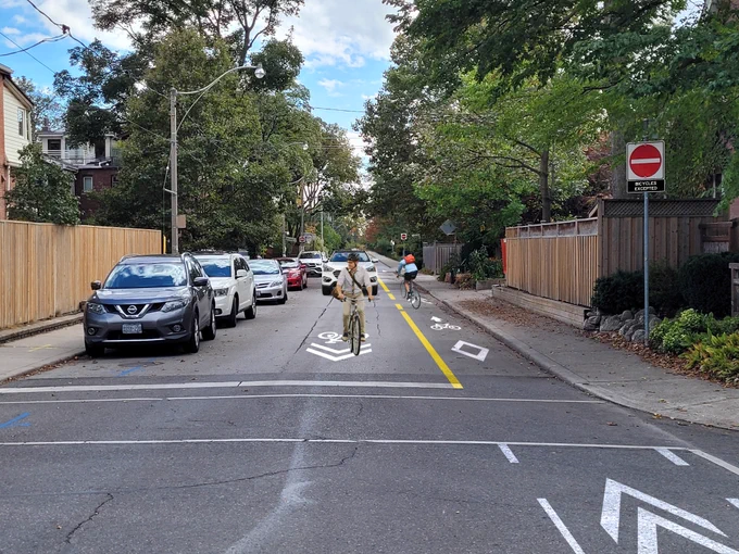 A rendering shows a neighbourhood greenway route, which includes a contra-flow bike lane, sharrows, and on-street parking. 