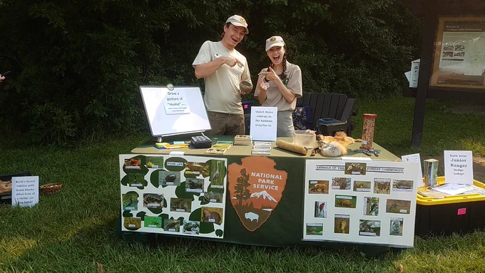 A young man and woman wearing volunteer uniforms stand behind a pop-up exhibit table by a park trailhead bulletin board.