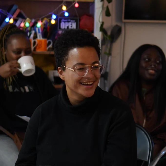 People sitting at YARD, smiling, laughing, drinking tea. The person in focus has short black curly hair, gold glasses, a big smile and a black turtle neck jumper.