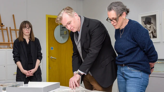 Two people look at items in a folder on a desk whilst another person looks on.