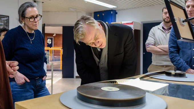 A man leaning down to look at a film reel set-up on a rewind desk, whilst other people look on.