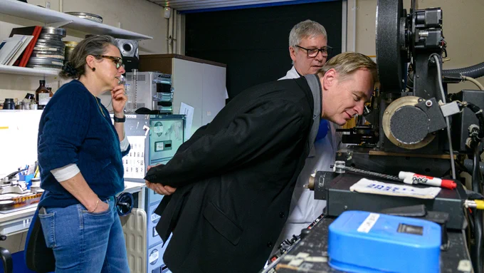 A man looking a microscope attached to a large machine holding a film reel. Two people look on.