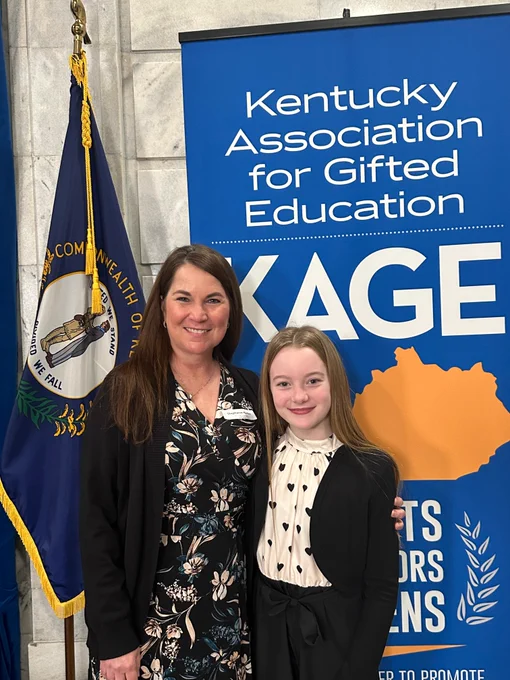Lydia and an adult pose for a picture in front of a banner that says "Kentucky Association for Gifted Education KAGE."