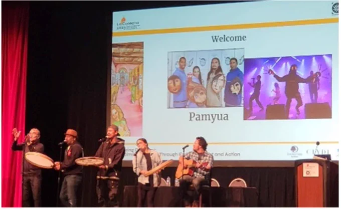 Five people are on a stage singing and playing instruments. Behind them is a screen with a welcome sign for the La Cosecha event.
