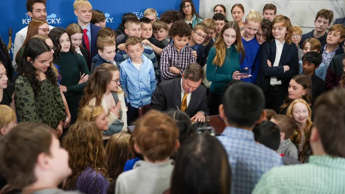 Gov. Andy Beshear writes on a paper with dozens of students standing around him and looking on.