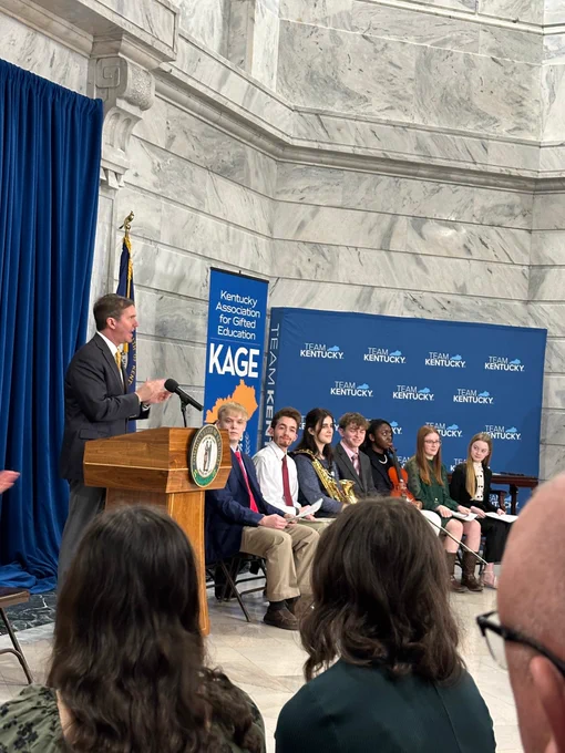 Gov. Andy Beshear speaks at a podium while seven individuals sit to the side of him.