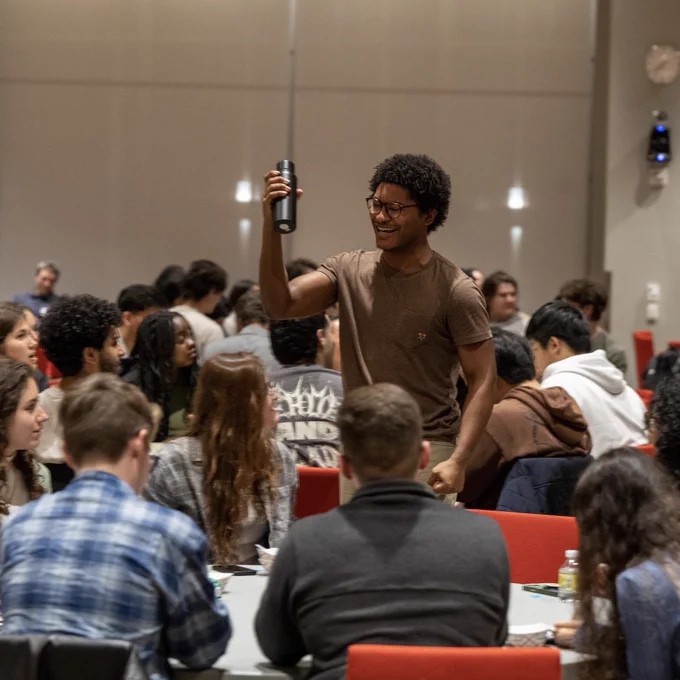 A young man stands up in a crowd of sitting people. He holds up a water bottle he won in a raffle.