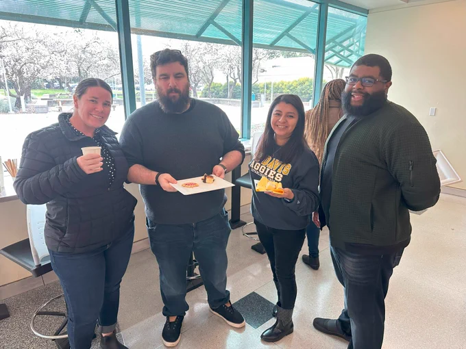 Four people stand indoors next to a window, some holding snacks
