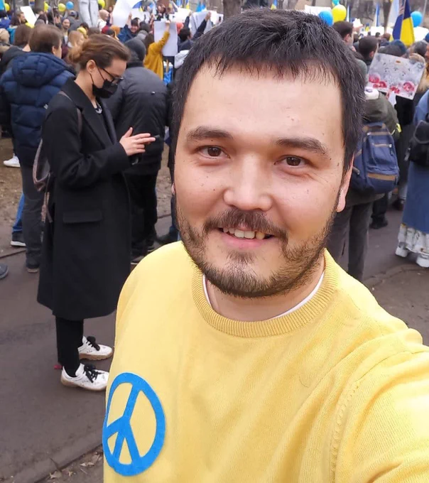 Human rights defender Aqylbek Muratbai taking a selfie while at a march in the streets. He is smiling and wearing a yellow sweater with the peace symbol on it.