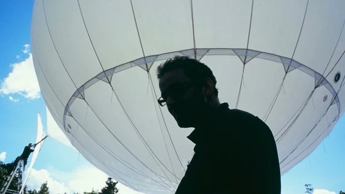 The silhouette of a person is seen in front of an enormous white airship taking up most of the image. A far away figure to the far left is standing on a ladder, working on the airship, and the scene is backdropped by a brilliant blue sky with white clouds.