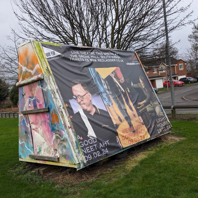 A giant banner on wooden frame on a village green advertises three plays