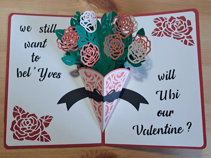 Une carte de saint valentin ouverte qui dévoile un bouquet de roses rouges et blanches découpées en relief. Autour du bouquet est écrit le texte "we still want to bel'yves, will Ubi our valentine ?"