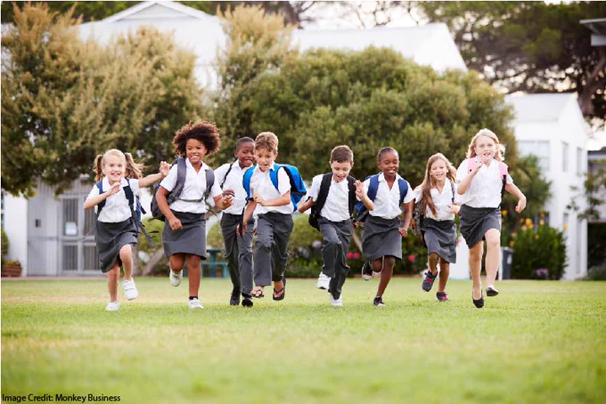 A group of young students -- all wearing white shirts and grey pants or skirts and backpacks -- racing across a lawn towards the camera