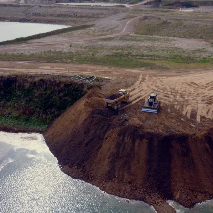 On a sheer cliff of soil surrounded by water, two heavy machines dig the earth.