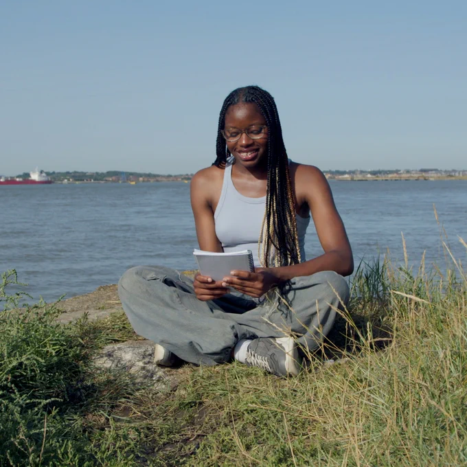 A person sits cross-legged on the grass banks of the river Thames, reading from a notebook in their hands.