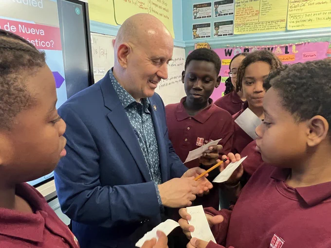 New York State Assembly Member, Harvey, Epstein, signs autographs on file cards for four 6th grade boys in  a classroom, standing in front of a SmartBoard. The students are wearing their PS 188 the Island School uniform shirts.