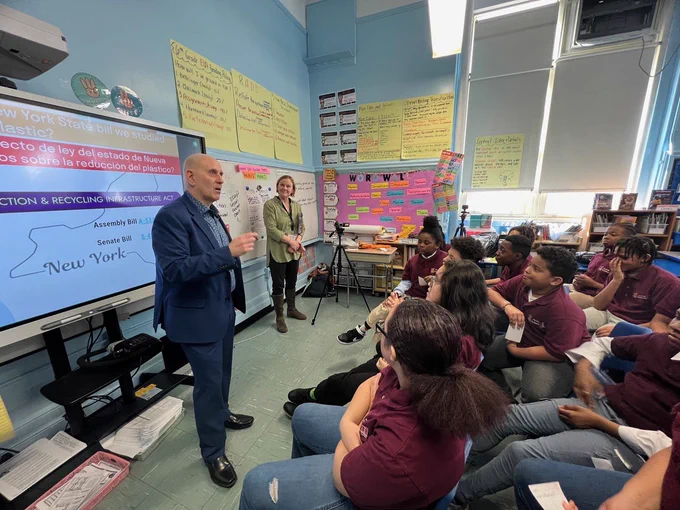 New York State Assembly Member, Harvey, Epstein, stands in front of a SmartBoard in a classroom, speaking to a students seated in chairs, listening to him speak.