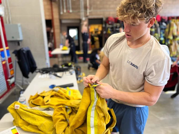 One 2024 SFF Recruit wearing an off-white shirt that says 'UC DAVIS' on it examining a wildland jacket.