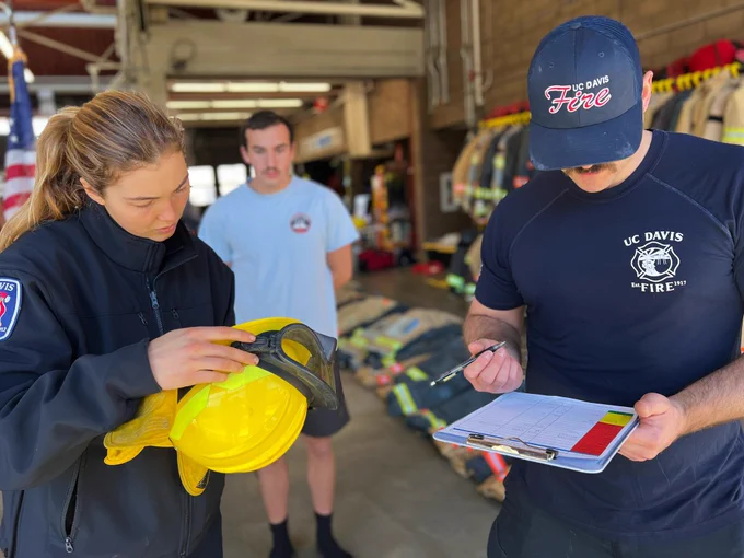 Firefighter M. Cullen and SFF Hallowell in navy blue UCDFD gear checking and marking off equipment requirements. One 2024 SFF Recruit with a white shirt in the background overseeing their actions.