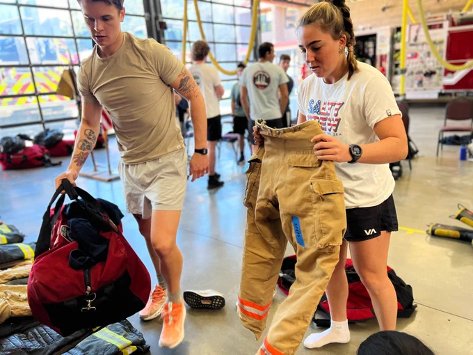 A 2024 SFF Recruit with a tan shirt and pumpkin-colored shoes carrying a red bag. Another 2024 SFF with a white shirt and black shorts carrying wildland pants.